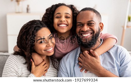 Love And Care. Portrait of cheerful African American family of three people hugging sitting on the sofa at home, posing for photo and looking at camera. Smiling young girl embracing her parents Royalty-Free Stock Photo #1927699334