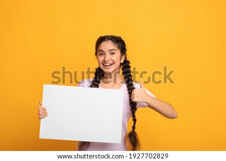 Place For Your Ad. Smiling indian woman holding empty blank board and showing thumbs up sign gesture isolated on yellow studio background. Happy lady standing with white square paper for template