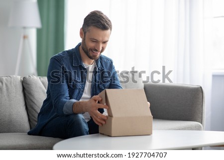 Happy middle-aged man unpacking delivery box, sitting on couch by tea table, home interior. Satisfied customer cheerful man opening package, ordering clothes or gadgets on Internet, copy space Royalty-Free Stock Photo #1927704707