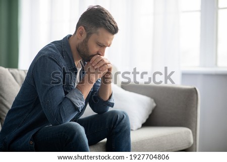 Sad bearded middle-aged man in casual sitting on couch at home, leaning on hands and looking down, upset man having problems, side view, copy space. Loneliness, depression, financial hangover concept Royalty-Free Stock Photo #1927704806