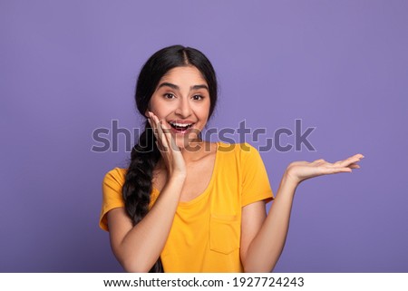 Wow, Look Here. Portarit of smiling young indian lady holding outstretched hand with open palm, advertising and showing free empty space and touching her cheek posing isolated on purple studio wall