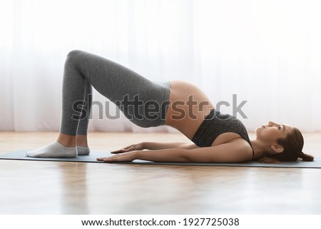 Prenatal Gymnastics. Young Pregnant Woman Training At Home, Doing Shoulder Bridge Exercise While Lying On Fitness Mat In Living Room, Expecting Lady Enjoying Healthy Lifestyle, Side View, Copy Space Royalty-Free Stock Photo #1927725038