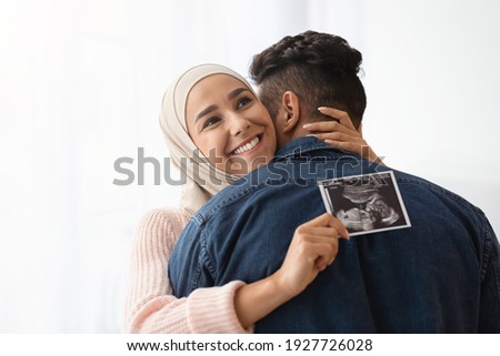 Expecting A Baby. Closeup Portrait Of Cheerful Pregnant Muslim Woman In Hijab Holding Ultrasound Picture And Hugging Her Husband, Standing Near Window At Home, Enjoying Healthy Pregnancy, Copy Space
