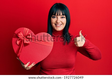 Young hispanic woman holding valentine gift smiling happy and positive, thumb up doing excellent and approval sign 