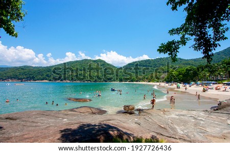 Beautiful seashore from brazilian coast of Sao Paulo. Crowded beach in a sunny Summer Day. People far away swiming and Sun bathing. Waves breaking into the sand. Happy people in vacation Royalty-Free Stock Photo #1927726436