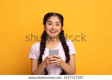 Amazing Application Concept. Portrait of happy smiling young indian woman using smartphone standing isolated on yellow studio background, looking at camera. Lady chatting online, browsing social media
