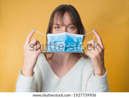 Portrait of a woman putting on a medical anti-virus mask. The photo was taken in a studio with a yellow background. The girl has blue eyes, blond hair and a white sweater.