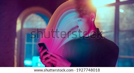 Cinematic portrait of stylish man in neon lighted interior using a smartphone. The face is smeared, sucked, absorbed into the screen. Concept of social network dependency, phone addiction.