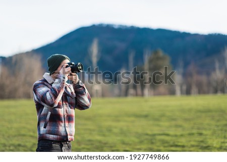 Man in winter clothes taking a photo standing in the middle of a field surrounded by mountains. Royalty-Free Stock Photo #1927749866