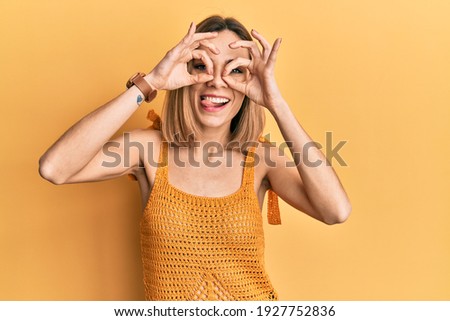 Young caucasian blonde woman wearing casual yellow t shirt doing ok gesture like binoculars sticking tongue out, eyes looking through fingers. crazy expression. 