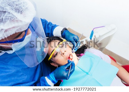 Latin American male dentist with bio safety suit attending and doing an oral examination to female patient. dental clinic, health and oral hygiene concept. Royalty-Free Stock Photo #1927772021