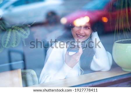 A European woman records and listens to voice messages in a new audio chat, a woman in headphones holds a smartphone in her hands, filmed through the glass of the cafeteria.
