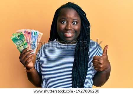 Young black woman with braids holding south african rand banknotes smiling happy and positive, thumb up doing excellent and approval sign 