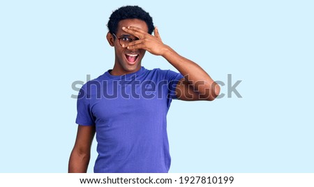 African handsome man wearing casual clothes and glasses peeking in shock covering face and eyes with hand, looking through fingers with embarrassed expression. 