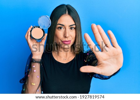 Young hispanic woman holding makeup powder with open hand doing stop sign with serious and confident expression, defense gesture 