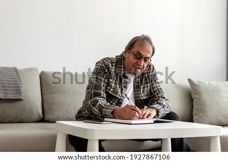 A senior Caucasian man in his 70s sitting at a table or desk, writing on a notepad at home. Senior grey hair man sitting in the living room and writing in his diary. Older man taking notes at home. 