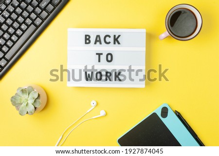 Back to work - text on display lightbox on yellow background workplace. Black keyboard, white earphones, coffee, tablet.