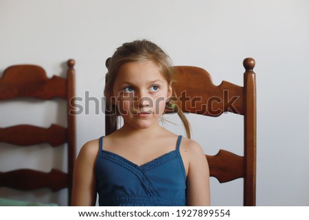 A girl of Slavic appearance sits at a table on a brown chair. against the background of a white wall. The girl is wearing a blue dress.м