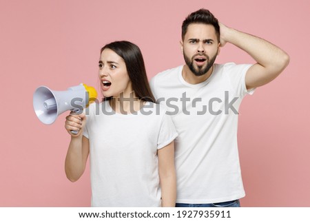 Young fun couple two friends man woman in white basic blank print design t-shirts hold scream in megaphone announces discounts sale Hurry up isolated on pastel pink color background studio portrait
