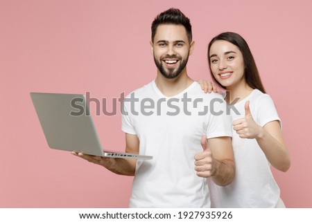 Young couple two friends man woman user in white basic blank print design t-shirts hold using work on laptop pc computer freelance remote jobs isolated on pastel pink color background studio portrait