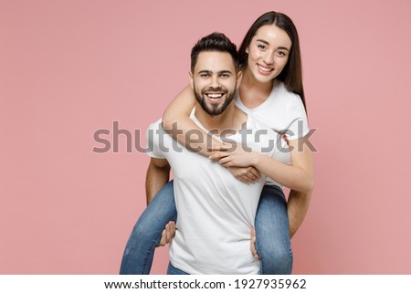 Young cheerful lovely couple two friends man woman in white basic blank t-shirts giving piggyback ride to joyful sitting on back fooling around isolated on pastel pink color background studio portrait