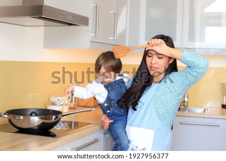 Young mother holding baby in her arms while cooking at modern kitchen home. Stressed and tired Asian woman with her kid. Housewife overwork concept. Royalty-Free Stock Photo #1927956377