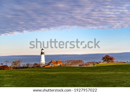 Lighthouse illuminated by the setting sun on a rocky promontory against the backdrop of a cloudy sky on the Atlantic coast in Portland Maine New England showing the ships a safe way to go