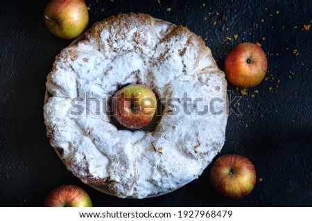 Round crust sprinkled with powdered sugar with a hole in the middle and apples on a dark background. Top view