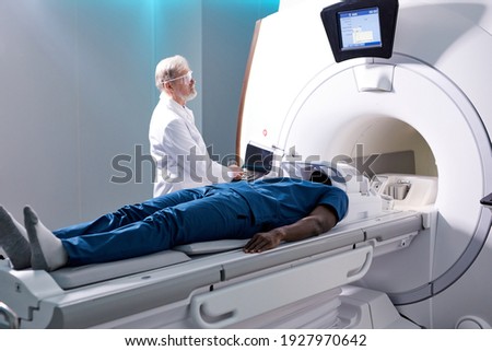 Senior Radiologist Controls MRI or CT or PET Scan with Male Patient Undergoing Procedure. High-Tech Modern Medical Equipment. Friendly Doctor Working With Patient Royalty-Free Stock Photo #1927970642