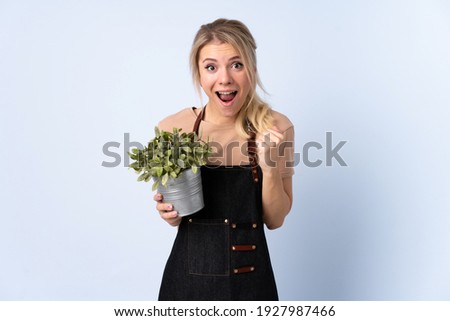 Blonde gardener woman holding a plant over isolated background celebrating a victory