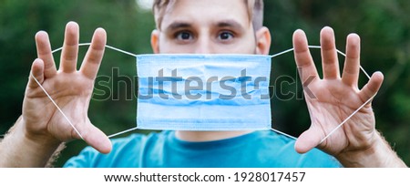 Scared Caucasian man holding blue medical mask in front of his face outside. Surgical disposable breathing mask as protection against coronavirus, epidemic covid 19 smog air pollution.Long web banner.