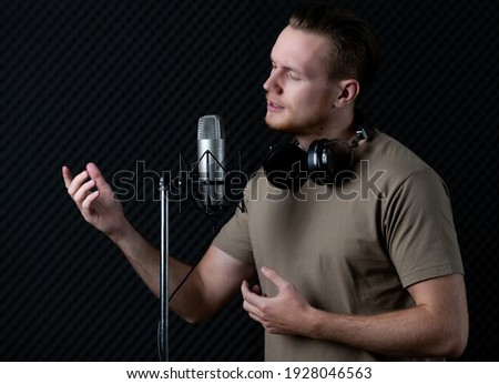 Young and handsome male singer working in voice recording studio, he sings to microphone with deep emotion and intention. Taken with studio light in dark style.
