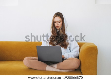 Young woman in white hoodie beautiful girl freelancer student using laptop sitting on yellow couch in the modern interior