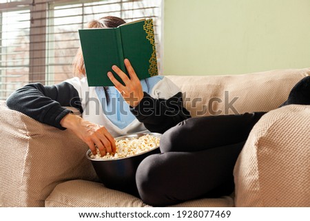 A young caucasian woman is lying with knees bent on an armchair and reading a green hard cover book by the window. Sunlight penetrates through the blinds. She nibbles on a bowl of popcorn as she sits.