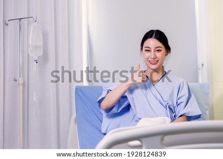 Portrait of a smiling Asian young female patient sitting on hospital bed room showing thumbs up gesture and sign to camera on recovery from sickness
