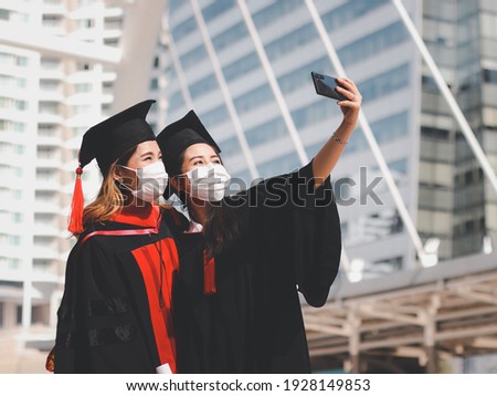 Two graduated women in graduation gowns and medical face masks,  making selfie photo with mobile phone. Class of 2021. Covid-19 prevention and new normal concept.