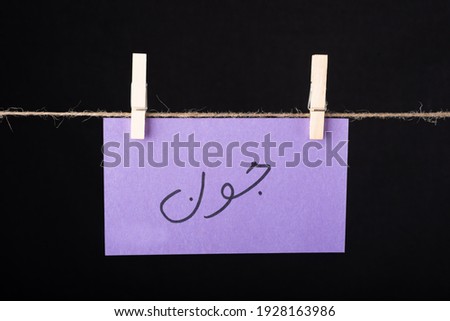
Translation June in urdu word written on a Purple color sticky note hanging with a wire on black background.