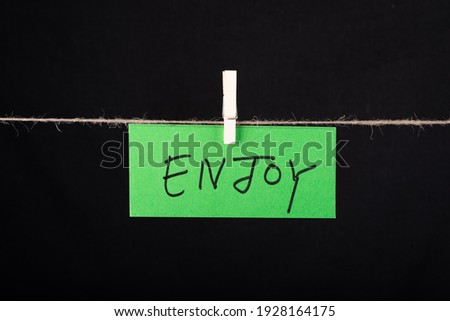 
Enjoy word written on a green color sticky note hanging with a wire on black background