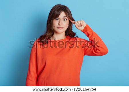 Portrait of dark haired woman in urban style jumper makes stupid sign with finger near head, gesturing bad mind, dumb insane idea, isolated on blue background, female looks at camera with astonishment