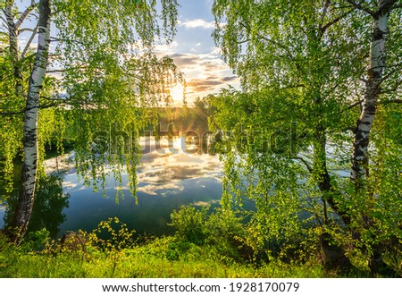 Scenic view at beautiful spring sunset on a shiny lake with green branches, birch trees, bushes, grass, golden sun rays, calm water ,deep blue cloudy sky and forest on a background, spring landscape Royalty-Free Stock Photo #1928170079