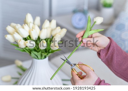 Cropped image of female florist cutting tulips flowers and putting in vase sitting at the living room coffee table. Composing bouquet. 