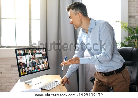 Successful male top manager or businessman dressed in formal stylish wear, standing near work desk in modern office, communicate with colleagues by video conference