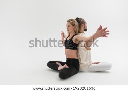 А man and a woman sit and meditate on the floor with their backs against each other. Young people practicing contact yoga psychological therapeutic exercise in studio on white background.