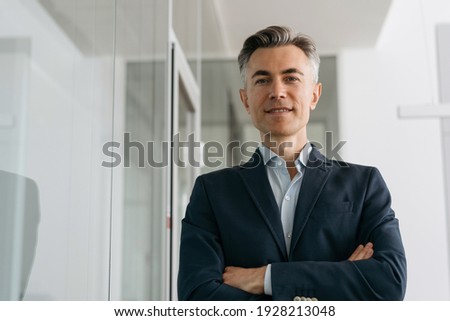 Confident mature business man with arms crossed looking at camera, smiling standing in modern office. Successful business concept  Royalty-Free Stock Photo #1928213048