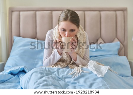 Sick young woman sitting at home in bed with hot cup of tea and handkerchief. Seasonal colds, cough, runny nose, viral infections, home treatment Royalty-Free Stock Photo #1928218934