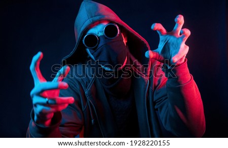 Scary guy in eyewear, mask and hood. Neon lighting. Young european man is in the dark studio.