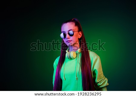 Photo of serious confident young woman dressed green pullover earphones dark glasses isolated vivid vibrant dark abstract background