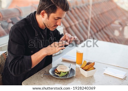 A young man taking picture with his smartphone of his meal - burger, roasted potato, beer on a terrace restaurant