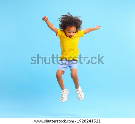 Full length of energetic little ethnic boy with Afro hairstyle in stylish casual outfit jumping with raised arms and looking down against blue background