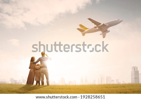 Back view of happy family embracing each other while standing at the park and looking at flying airplane in dusk sky